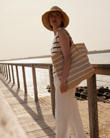Woman standing on a wooden dock by the water, holding a striped bag.