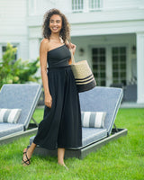 Model laughing on a lawn wearing black organic cotton beach pants with elastic waistband with drawstring and tassel detail with a black one piece one shoulder bathing suit holding a black and tan beach bag