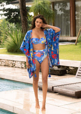 Woman in a colorful bikini and kimono standing by a pool with greenery and a building in the background.