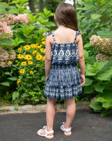 Young girl in a blue patterned dress standing in front of a garden with flowers and greenery.