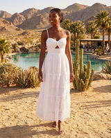Woman in a white dress standing in a desert landscape with mountains and a pool in the background.