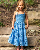 Young girl in a blue dress standing outdoors on stone steps with greenery in the background