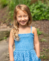 Young girl in a blue dress standing outdoors with greenery in the background