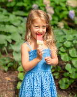 Young girl in a blue dress eating a popsicle outdoors with greenery in the background
