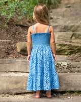 Young girl in a blue dress standing on a rocky path with greenery.