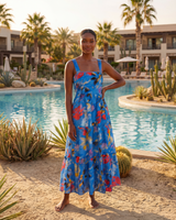 Woman in a blue floral dress standing by a pool with palm trees and buildings in the background.