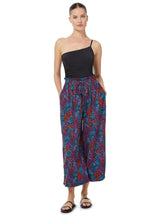 The back of a model wearing a floral pattern organic cotton beach pants with elastic waistband with drawstring and tassel detail with one piece black bathing suit with one shoulder