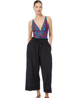 Model posing with her hands in her pocket wearing black organic cotton beach pants with elastic waistband with drawstring and tassel detail with a floral deep plunge one piece bathing suit