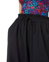 Model posing with her hands in her pocket wearing black organic cotton beach pants with elastic waistband with drawstring and tassel detail with a floral deep plunge one piece bathing suit