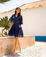 Woman in a navy dress standing outdoors with a palm tree and white wall in the background