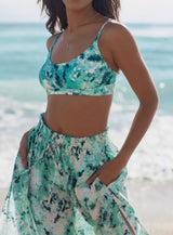 Model standing in front of ocean wearing an ocean print scoop neckline bikini top with under-bust band and adjustable straps with matching skirt