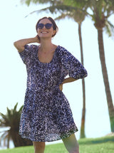 model laughing and standing in front of palm trees with her hand behind her neck wearing a black and white leopard print slightly above the knee dress with rounded neckline and short puff sleeve with a covered elastic cuff with black sunglasses