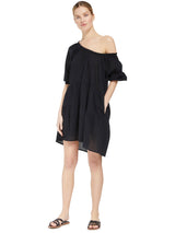 model posing with her hand in her pocket wearing a black slightly above the knee dress with rounded neckline and short puff sleeve with a covered elastic cuff with black sandals and wearing the dress off one shoulder
