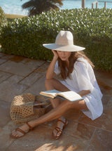 model sitting on the floor reading a book in front of the beach wearing a white slightly above the knee dress with rounded neckline and short puff sleeve with a covered elastic cuff and accessorized with a straw hat, crochet bag, and brown sandals