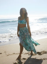 Model standing in front of ocean wearing an ocean print scoop neckline bikini top with under-bust band and adjustable straps with matching skirt
