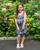 Young girl in a blue dress standing in a garden with flowers and greenery.