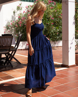A woman wearing a navy blue tiered maxi dress with spaghetti straps, standing on a tiled patio under a white pergola with green foliage in the background.