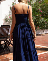 A woman viewed from behind wearing a dark navy blue maxi dress with thin spaghetti straps, a fitted bodice, and a tiered skirt, standing on a tiled patio with green foliage in the background.