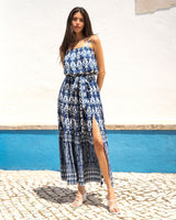 Woman wearing a blue and white patterned dress standing on a cobbled street with a white wall and blue baseboard in the background.