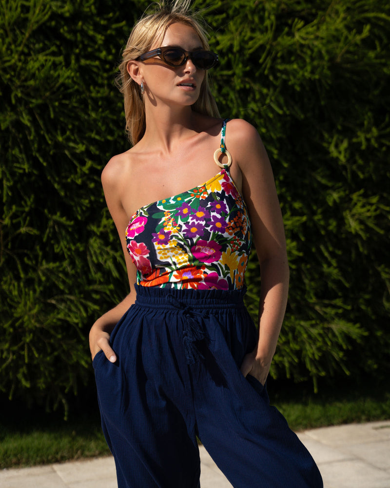 Woman wearing a colorful floral top and navy pants against a green hedge.
