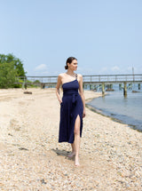 Model with head turned towards ocean wearing navy one shoulder strap one piece bathing suit