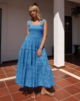 Woman in a blue dress standing in a sunlit indoor courtyard with a tiled floor and white walls.