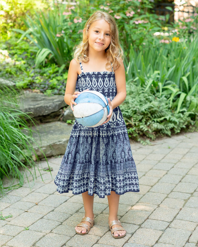 Young girl in a blue patterned dress holding a blue and white ball in a garden setting.