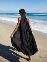 The back of model walking on a beach wearing an unbuttoned black sleeveless, high neck with ruffle detail,  shirt dress.