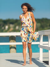 Brunette model with curly hair walks on a white dock wearing a Floral Ikat print dress with an elastic waistband, adjustable black drawstring tassels and a V-notch neckline with a ruffled skirt and pockets.