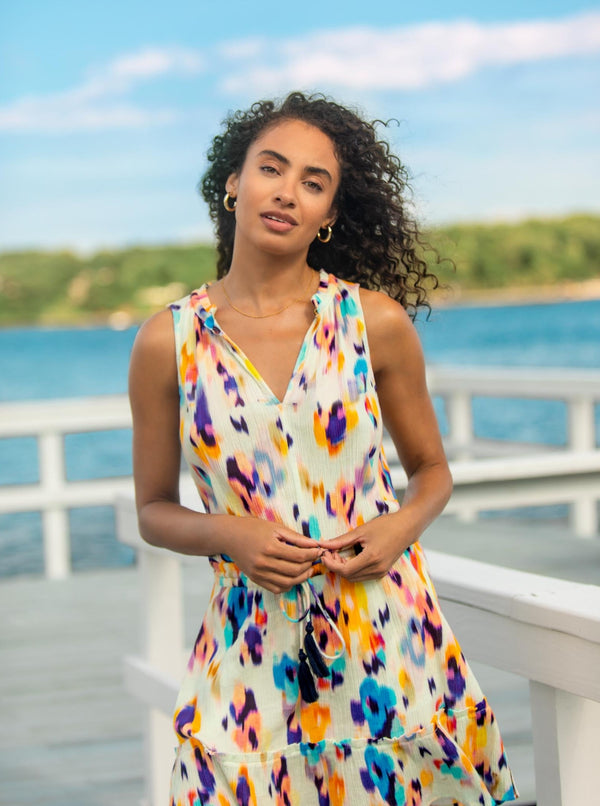 Brunette model with curly hair posses on a white dock wearing a Floral Ikat print dress with an elastic waistband, adjustable black drawstring tassels and a V-notch neckline with a ruffled skirt and pockets.
