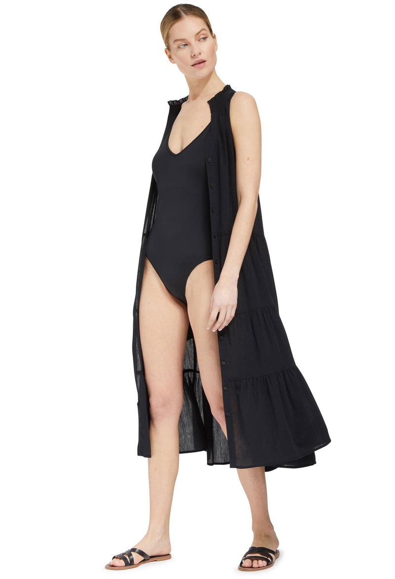 Model looking to the side wearing an unbuttoned black sleeveless, high neck with ruffle detail,  shirt dress with a back one piece bathing suit. 