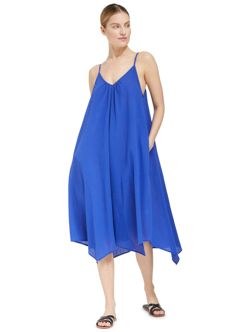 Model posing with hands in her pockets wearing a cobalt blue strappy and long flowy dress with adjustable back shoulder straps, and v-neckline front and back with black sandals