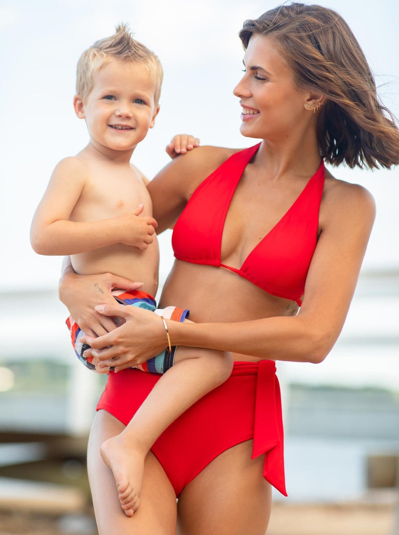 Model holding a toddler wearing a cherry red bikini top with halter neck tie, adjustable cups, and a spaghetti tie under the bust with matching high waist bikini bottoms with a side tie 