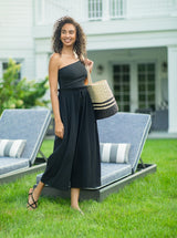 Model laughing on a lawn wearing black organic cotton beach pants with elastic waistband with drawstring and tassel detail with a black one piece one shoulder bathing suit holding a black and tan beach bag 