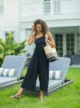 Model laughing on a lawn wearing black organic cotton beach pants with elastic waistband with drawstring and tassel detail with a black one piece one shoulder bathing suit holding a black and tan beach bag 