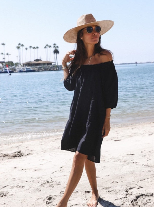 girl standing on the beach wearing a black slightly above the knee dress with rounded neckline and short puff sleeve with a covered elastic cuff with a straw hat and round sunglasses  