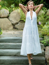 Model wearing fresh white sleeveless, high neck with ruffle detail, buttoned shirt dress with optional matching belt with tassels and pockets with hands behind her neck. 