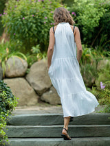 The back of model walking up concrete stairs wearing a fresh white sleeveless, high neck with ruffle detail, buttoned shirt dress with hands in pockets. 