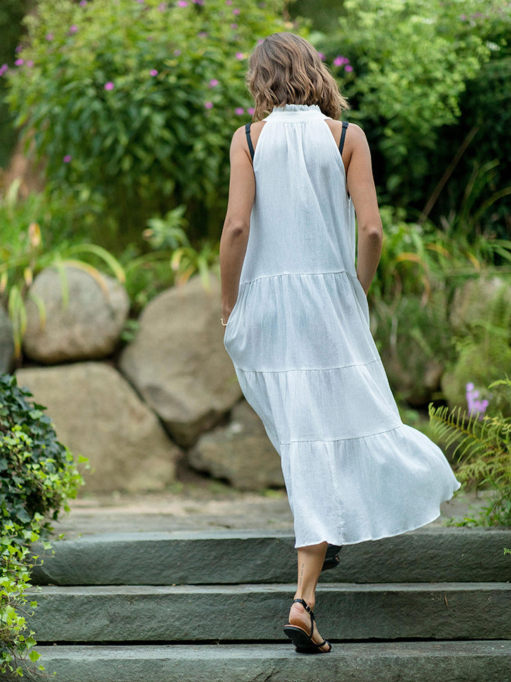 The back of model walking up concrete stairs wearing a fresh white sleeveless, high neck with ruffle detail, buttoned shirt dress with hands in pockets. 
