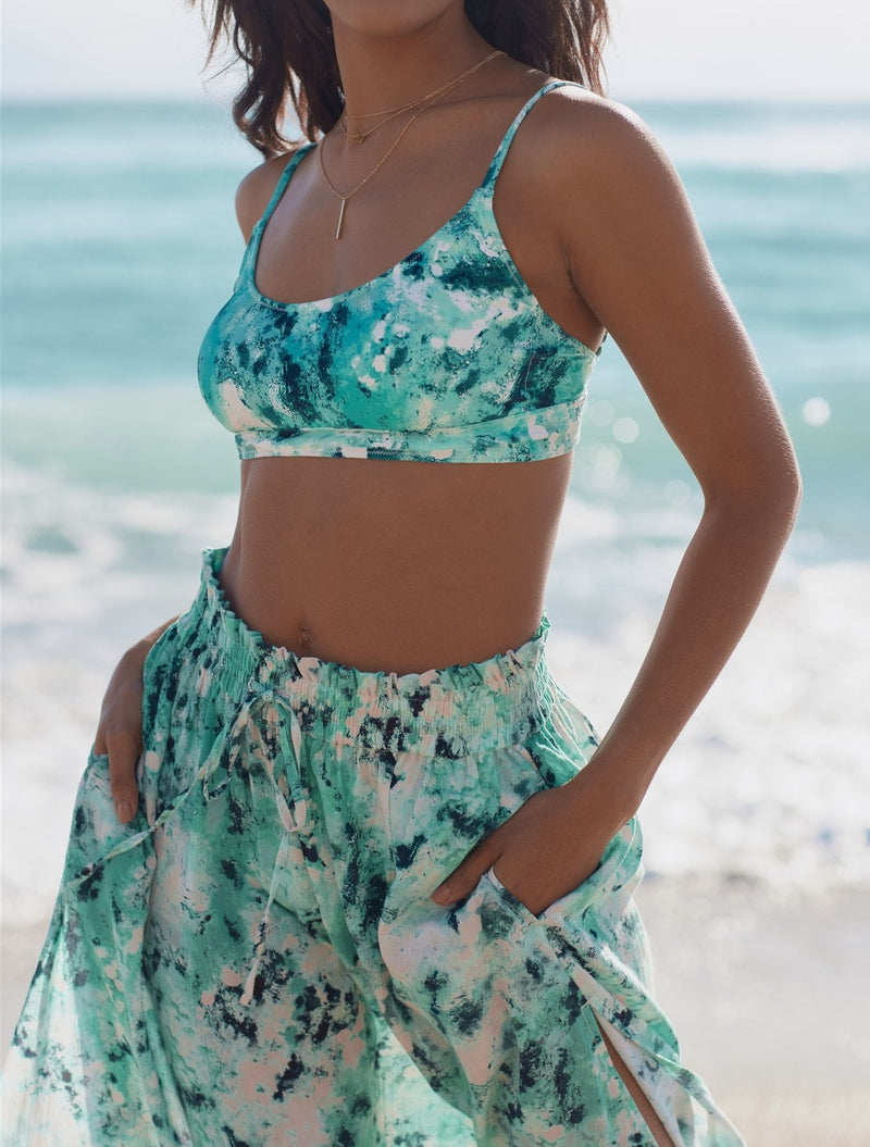 Close up shot of model walking on beach with hands in her pockets wearing ocean print inspired midi length skirt with side slits and elastic waistband with drawstring detail  with matching ocean print bikini top and layered gold necklaces 