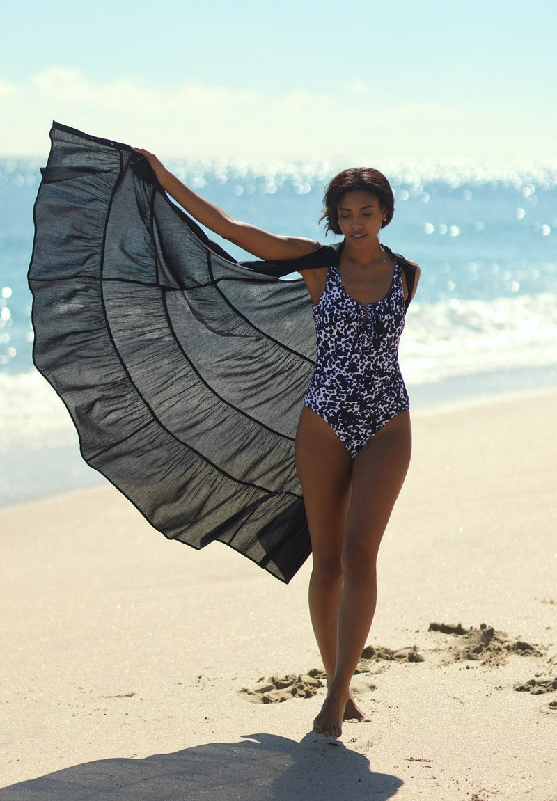 Model walking on the beach looking down wearing an unbuttoned black sleeveless, high neck with ruffle detail,  shirt dress with a black and white leopard one piece bathing suit. 