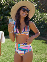 Model laughing outside in the yard in a geometric and graphic colored plunge neckline, classic bikini top with adjustable straps and an elastic underband with a matching classic midrise bikini bottom and a straw hat, sunglasses, and wine glass