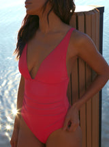 Model in front of a lake leaning on a wooden beam wearing a french cup coral red one piece swimsuit with a deep plunge-neckline, adjustable back straps, and shirring at the front waist with power compression mesh lining at the front lower torso