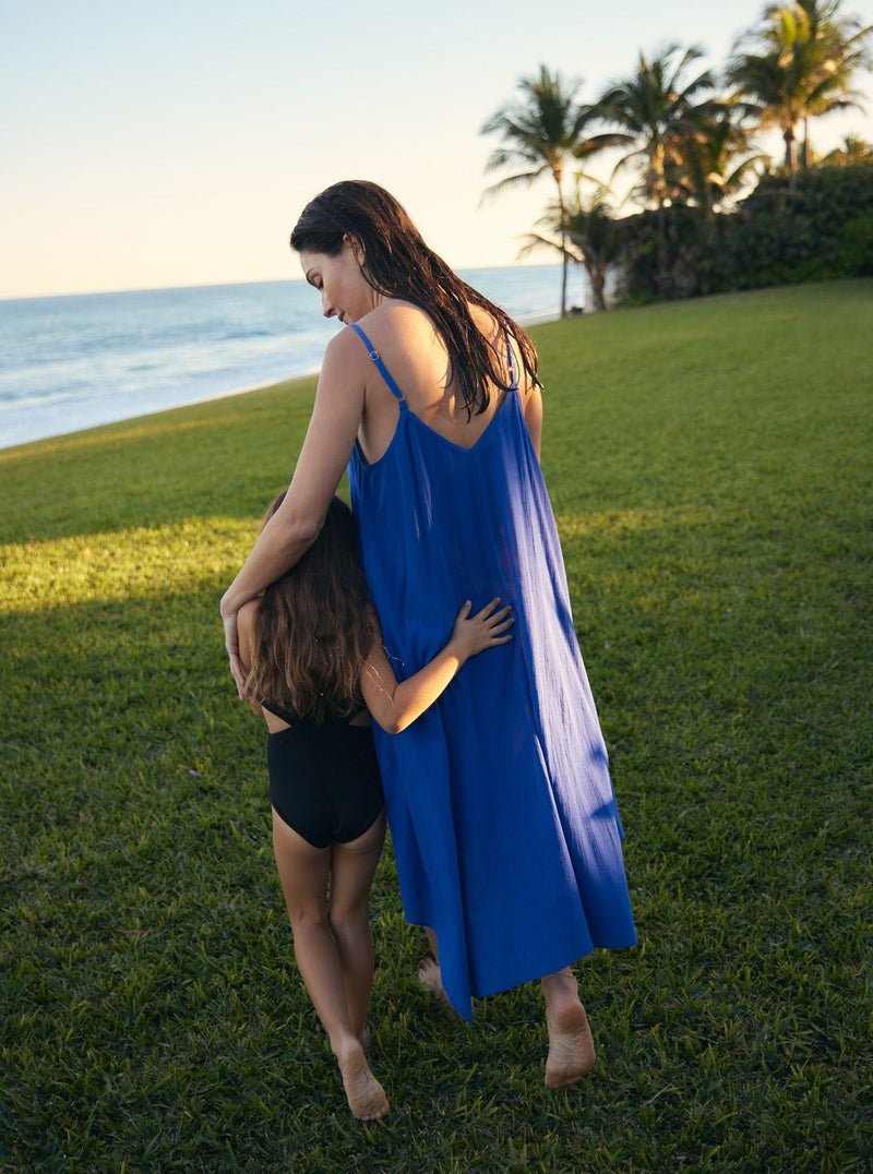 Model wearing a cobalt blue strappy and long flowy dress with adjustable back shoulder straps, and v-neckline front and back walking side by side with little girl in front of the beach