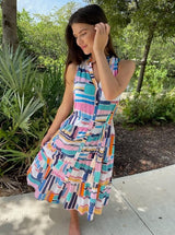 Model grinning and putting her hair behind her ear wearing a geometric and graphic colored sleeveless, high neck with ruffle detail, buttoned shirt dress with optional matching belt with tassels and pockets. 
