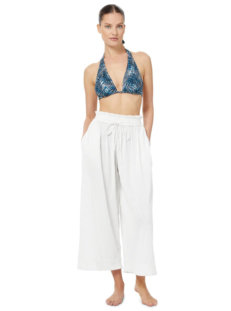 Model posing with her hands in her pockets wearing white organic cotton beach pants with elastic waistband with drawstring and tassel detail with blue and white patterned triangle bikini top 