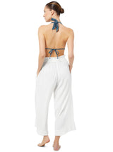 The back of a model posing with her hands in her pockets wearing white organic cotton beach pants with elastic waistband with drawstring and tassel detail with blue and white patterned triangle bikini top 