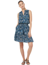 Model posses in front of white background wearing black leather sandals and a cotton dress with ruffles around the neck and a v-neck cut. The dress has a elastic waist with table ties hanging down from the waist. Dress is in a teal and white wave print.   