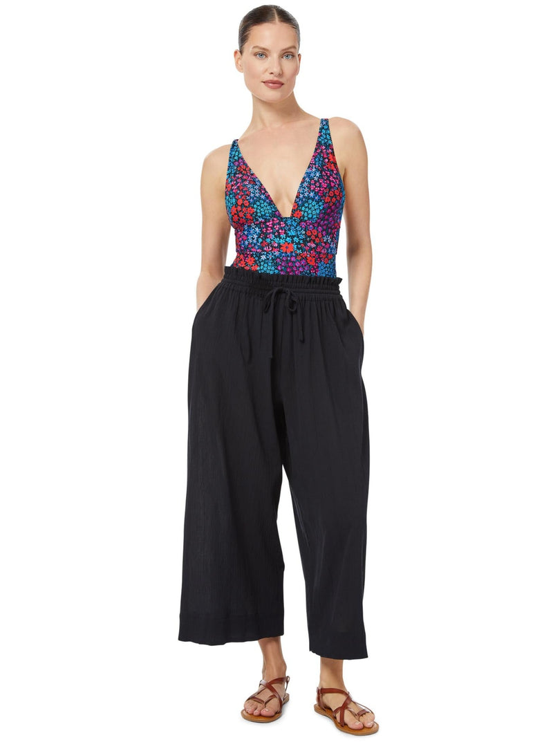 Model posing with her hands in her pocket wearing black organic cotton beach pants with elastic waistband with drawstring and tassel detail with a floral deep plunge one piece bathing suit