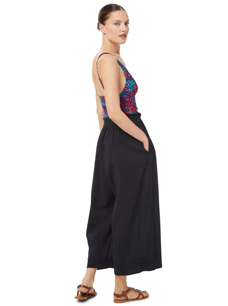 The back of a model wearing  black organic cotton beach pants with elastic waistband with drawstring and tassel detail with a floral deep plunge one piece bathing suit